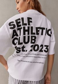 Bijela prevelika majica s podebljanim crnim tekstom koji glasi „SELF ATHLETIC CLUB est. 2023.“ Pamučni materijal s ležernim krojem.