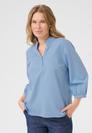 Lichtblauwe blouse met een mandarijnenkraag, drie knopen en poffende mouwen. Gladde stof, relaxte pasvorm en zijzakken.
