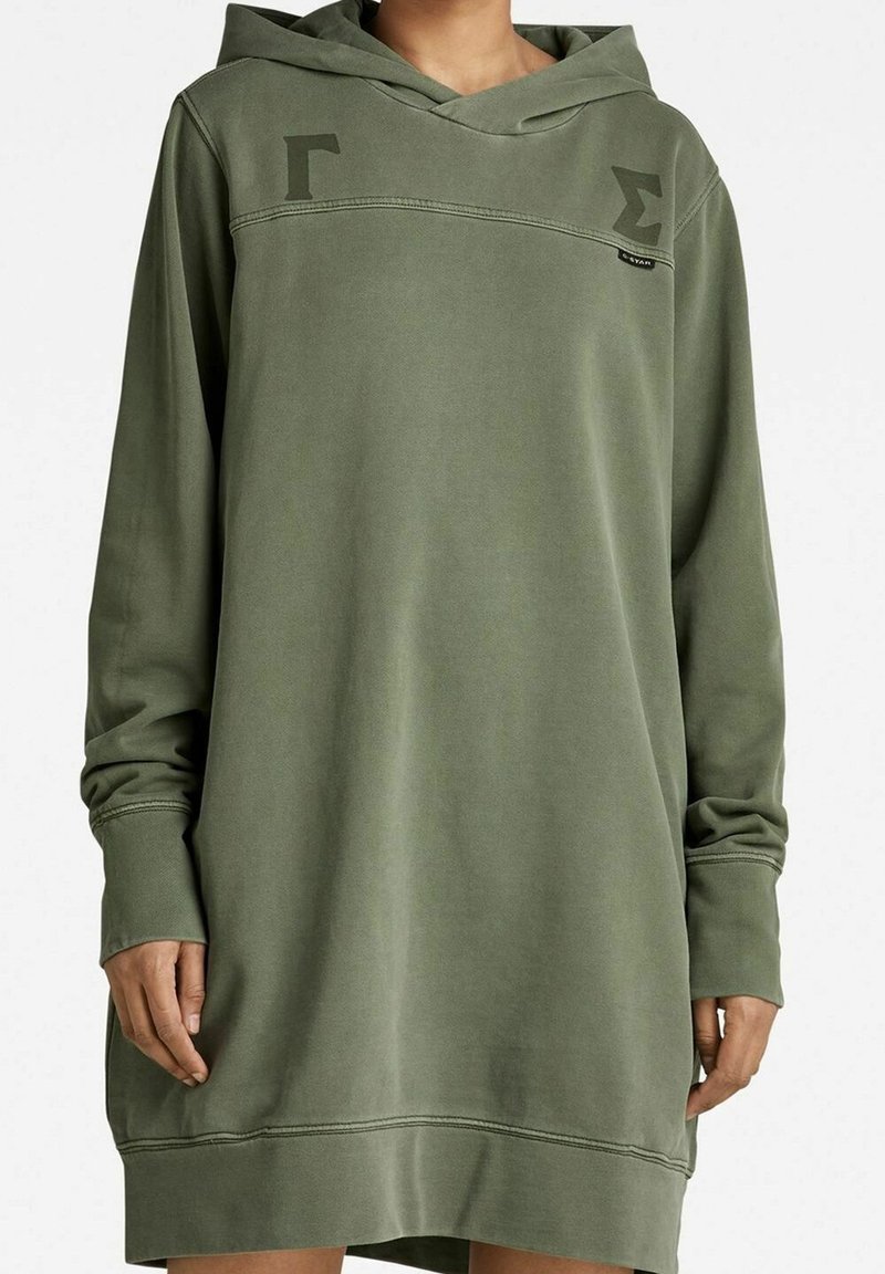 Oliivinvihreä hupparimekko, joka on valmistettu pehmeästä kankaasta. Siinä on suuri huppu, pitkät hihat ja hienovaraiset graafiset yksityiskohdat edessä. Rintakehällä kulkee vino sauma.