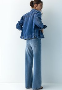 Frau steht mit Blick nach links, trägt eine Jeansjacke und hochtaillierte, weit geschnittene Jeans vor einem schlichten weißen Hintergrund.