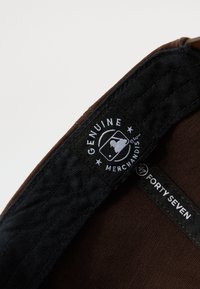 Brun stoff baseball caps innside som viser en svart etikett med "Genuine Merchandise" og "Forty Seven" i hvit tekst. Teksturert fôr synlig.
