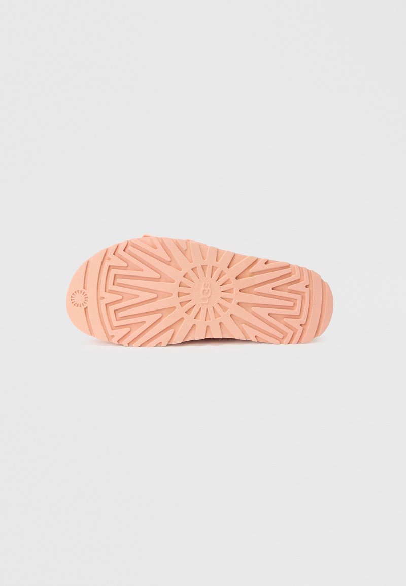 Gummisohlen in hellpfirsichfarbener Ausführung, mit einer strukturierten Oberfläche, die geometrische Muster und einen kreisförmigen Akzent an der Ferse aufweist.