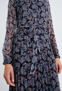 Φόρεμα μακρύ μανίκι από διάφανο μαύρο ύφασμα με πολύχρωμο σχέδιο paisley, με σφηνοειδή μέση και ζώνη, και υφασμάτινα μανίκια.