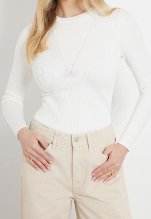 Femme portant un haut blanc à manches longues côtelé avec des détails tricotés géométriques et un pantalon beige taille haute, les mains dans les poches.
