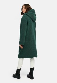 Manteau en polaire verte avec une capuche, présentant une texture douce, des manches longues et un design mi-long. Porté sur un pantalon blanc et des bottes noires.