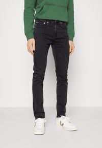 Jeans de mezclilla negros con corte ajustado, que presentan estilo de cinco bolsillos y herraje plateado. Combinados con una sudadera verde y zapatillas blancas.