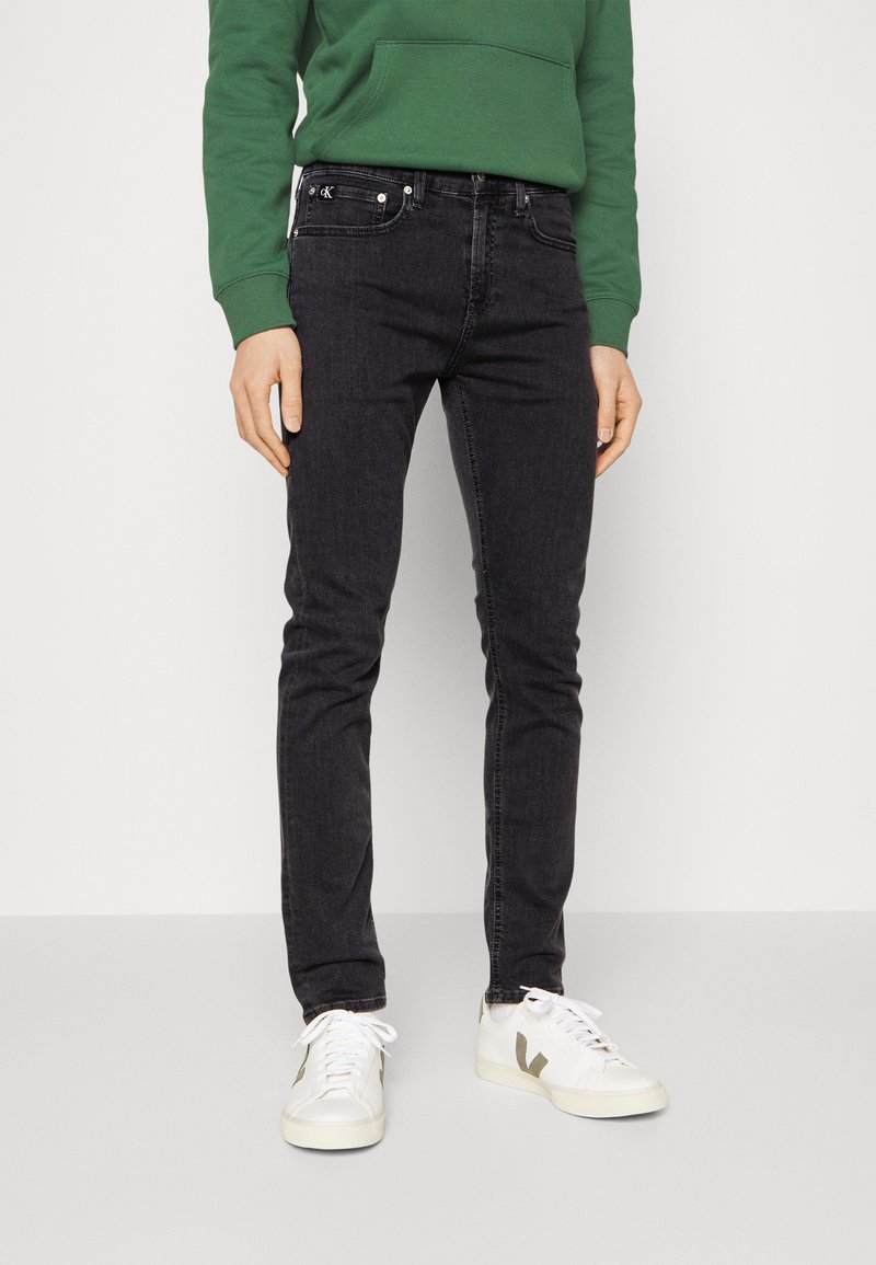 Jeans de mezclilla negros con corte ajustado, que presentan estilo de cinco bolsillos y herraje plateado. Combinados con una sudadera verde y zapatillas blancas.