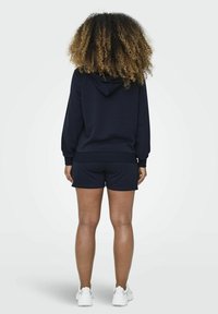 Felpa blu navy con cappuccio e pantaloni corti coordinati. Tessuto morbido, vestibilità rilassata, polsini e orlo a costine. Sneakers bianche completano l'outfit.