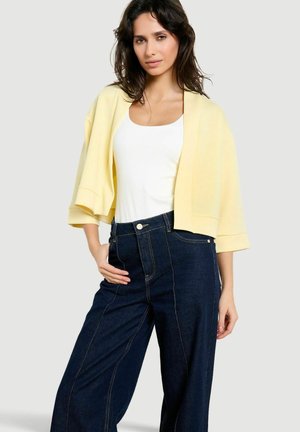 Donna che indossa una bolero giallo pallido sopra una canotta bianca e jeans a vita alta a gamba larga di colore blu scuro, in piedi con una mano sui fianchi.