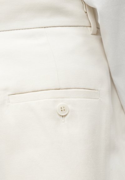 Gros plan sur un pantalon beige montrant une poche arrière avec un bouton et une boucle de ceinture sur un tissu lisse.