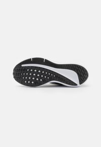 Semelle de chaussure de sport noire et blanche avec des motifs de crampons et des rainures pour l’adhérence sur fond blanc uni.