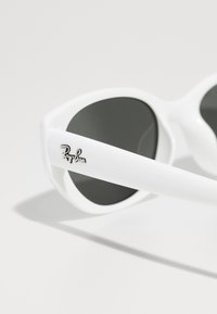 Bijele sunčane naočale s okruglim staklima, crnim zatamnjenim staklima i glatkim plastičnim okvirom. Imaju srebrni Ray-Ban logo sa strane.