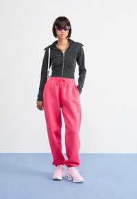 Čierna bunda na zips, ružové tepláky s príťahom a ružové tenisky. Elegantný dizajn s logom na teplákoch, na svetlom pozadí.
