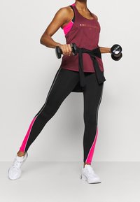 Mörkröd linne med svarta accenter och logotyp, i kombination med svarta leggings med rosa sidopaneler och vita ränder, hållande hantlar.