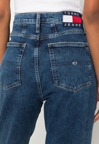 Donkere blauwe spijkerbroek met een hoge taille, voorzien van twee achterzakken en een logopatch in rood, wit en blauw op de tailleband.