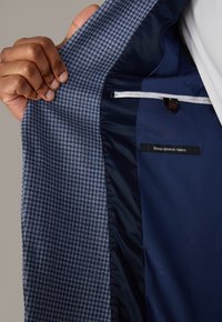 Wnętrze dopasowanej marynarki z niebiesko-białym wzorem pepitki, gładką granatową podszewką, czerwonym przeszyciem akcentującym oraz metką z napisem "najlepsza tkanina elastyczna".