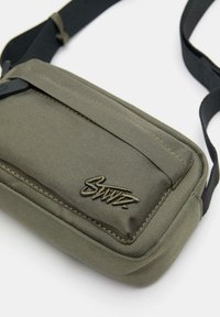 Zielona oliwkowa torba na ramię z materiału, z zamykaną na zamek kieszenią i widocznym haftowanym logo. Posiada czarny pasek i teksturowaną powierzchnię.