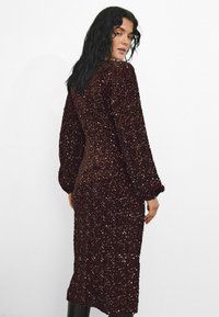 Bronzas krāsas sequins kleita ar garām piedurknēm, pieguļoša silueta un teksturētām rotājumām, sasniedzot vidusslavu. Zemāk ir gluds audums.