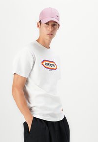 Hvid bomulds t-shirt med et farverigt "RIPCURL" logo, parret med sorte shorts. En lyserød kasket med et logo fuldender looket.