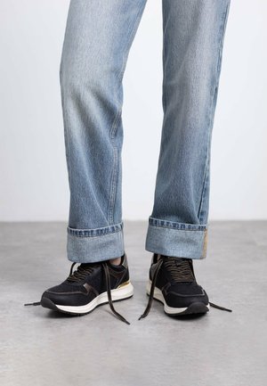 Zapatillas deportivas negras con tejido texturizado, detalles en dorado y suelas de color claro. Llevadas con jeans de mezclilla azul doblados.