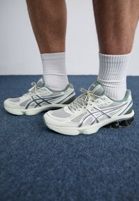 Ténis cinza claro com detalhes em prata, parte superior em malha, sola curva e almofada de gel preta. Combinados com meias brancas caneladas, sobre um tapete azul.