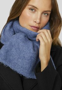 Blå, tekstureret uldscarf viklet om halsen, med en blød finish og frynser, båret sammen med et sort ydre tøj.