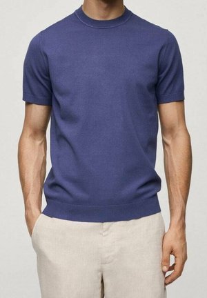 Hombre con un jersey azul ajustado de manga corta y cuello redondo, con pantalones beige, de pie con una mano en el bolsillo frente a un fondo liso.