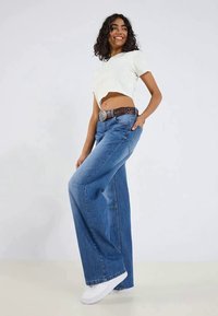Top curto verde claro, jeans azuis de corte relaxado com pernas largas e tênis brancos. Cinto castanho com detalhe decorativo na fivela.
