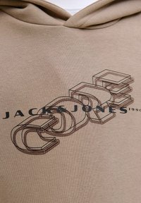 Sudadera beige con un gran estampado gráfico que presenta "JACK & JONES" en texto negro y la palabra "CORE" en estilo 3D, contorneada en tonos más oscuros.