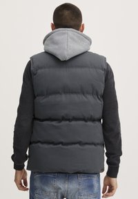 Ermeløs grå dunstvest med kviltet design, båret over en grå hettegenser. Den har ribbestrikket krage og fullførte ermehull.