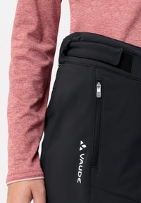 Zwarte buitenbroek met een ritszak, klittenbandsluiting en wit VAUDE-logo. Gecombineerd met een lange mouwen top in rood gemêleerd.
