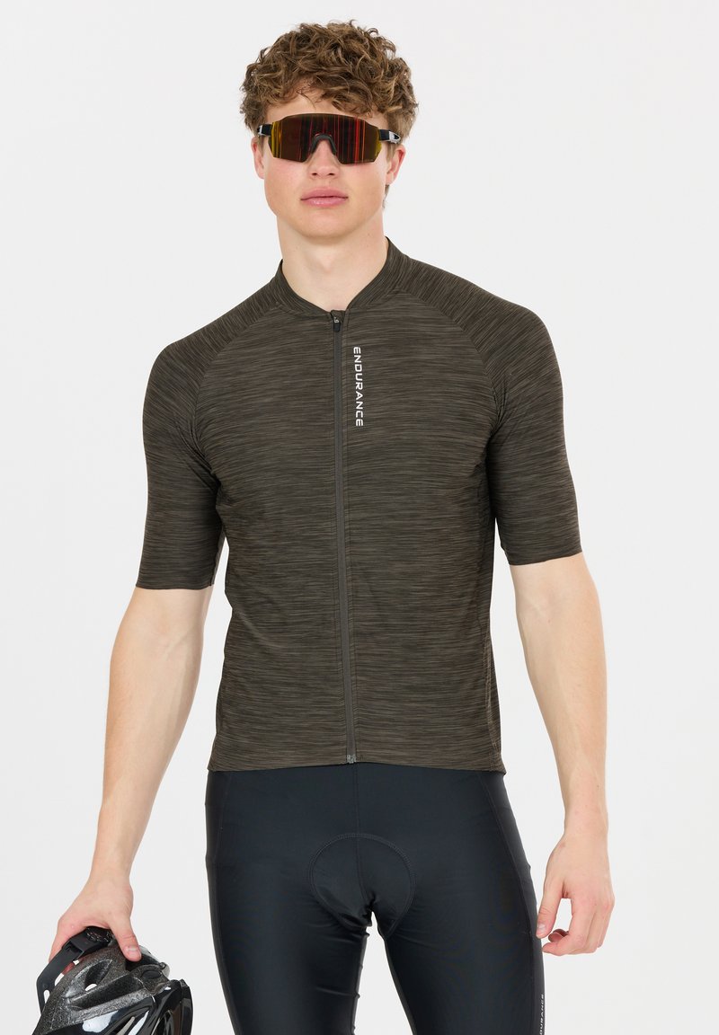 Jeune homme portant un maillot de cyclisme foncé et un short, tenant un casque de vélo et portant des lunettes de sport réfléchissantes.