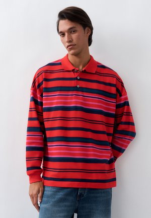 Νεαρός άνδρας με κόκκινο πόλο με μακριά μανίκια, με οριζόντιες ροζ, ναυτίλια και πράσινες ρίγες, στέκεται μπροστά σε έναν λευκό τοίχο.