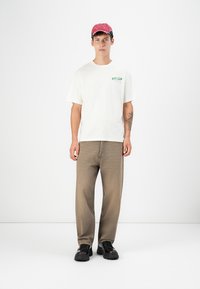 White cotton t-shirt with green logo, beige denim pants, and a red baseball cap. Chunky black shoes complete the outfit.