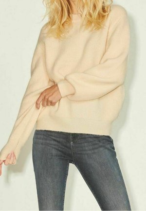Kobieta o falowanych blond włosach, ubrana w beżowy, oversize'owy sweter oraz ciemnoszare dopasowane jeansy, poprawiająca rękaw swetra.