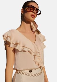 Light pink blouse with ruffled sleeves, textured fabric, paired with a gold chain belt and oversized tortoise-shell sunglasses.