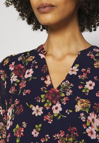 Blouse marine à fleurs avec un col en V, présentant une variété de motifs floraux roses, rouges et verts sur un tissu doux.