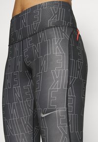 Zbliżenie czarnych legginsów Nike z białym konturze tekstowym logo i małym odblaskowym swooshem na udzie.