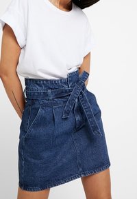 Denim-Minikleid mit taillierter Taille, Fronttaschen und leicht abgerundetem Saum. Der Stoff hat eine mittlere blaue Waschung und eine weiche Textur.