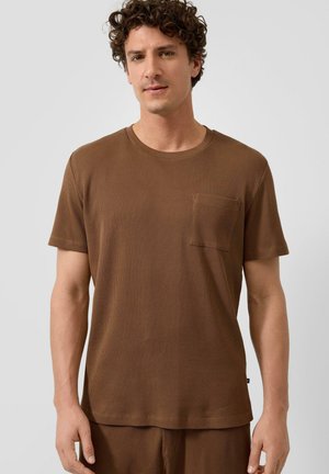 Mężczyzna o kręconych włosach, ubrany w brązową teksturowaną koszulę z krótkim rękawem i kieszenią na piersi oraz dopasowane spodnie na jednolitym tle.