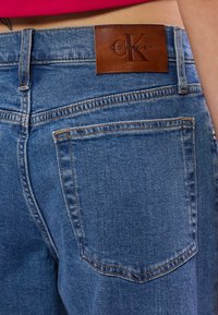 Jeans em denim em um tom azul claro, com bolsos traseiros, costura visível e um patch de couro castanho estampado com "Calvin Klein" na cintura.