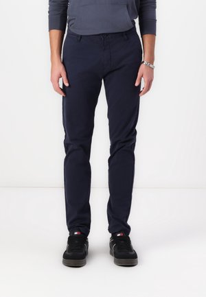 Personne portant un pantalon slim bleu marine foncé, des baskets noires avec un logo rouge et blanc, et un haut à manches longues foncé, debout sur un fond blanc.