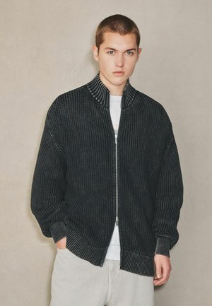 Junger Mann mit kurzen Haaren, trägt einen schwarzen, gerippten Reißverschluss-Pullover über einem weißen Hemd und hellfarbigen Hosen, steht vor einem schlichten Hintergrund.