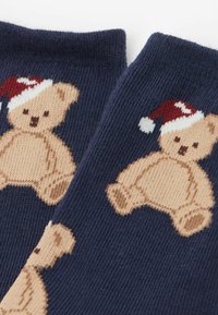Marineblå sokker med beige teddy-bear designs, iført røde og hvide hatte. Strikket tekstur med legende mønstre og blødt stof.