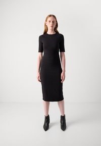 Crna bodycon haljina s kratkim rukavima, okruglim izrezom i dužinom do koljena. Glatka tkanina s minimalnim detaljima. Nosena s čizmama do članka.