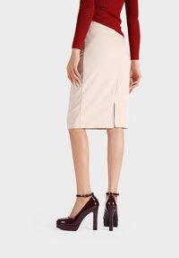 Décolleté in vernice bordeaux con cinturino alla caviglia, caratterizzate da una punta tonda e una soletta, indossate con una gonna a tubino beige e una maglietta rossa.