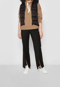 Gilet imbottito nero sopra un hoodie beige, abbinato a pantaloni neri con fessure laterali e sneaker bianche. Texture lisce e design contemporaneo.