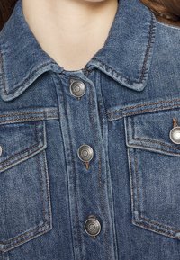 Chaqueta de denim con cuello clásico, botones a presión plateados, dos bolsillos en el pecho y costuras en contraste en un lavado azul medio.