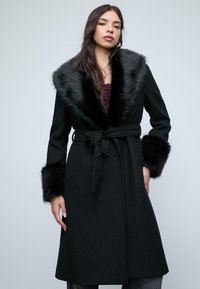 Giovane donna che indossa un cappotto nero con cintura, colletto di pelliccia grande e polsini di pelliccia, in piedi davanti a uno sfondo grigio chiaro uniforme.