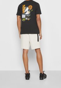 Man med svart Nike t-shirt med grafik av solros och vita shorts, stående vänd mot en vägg, svarta sneakers, enkel grå bakgrund.